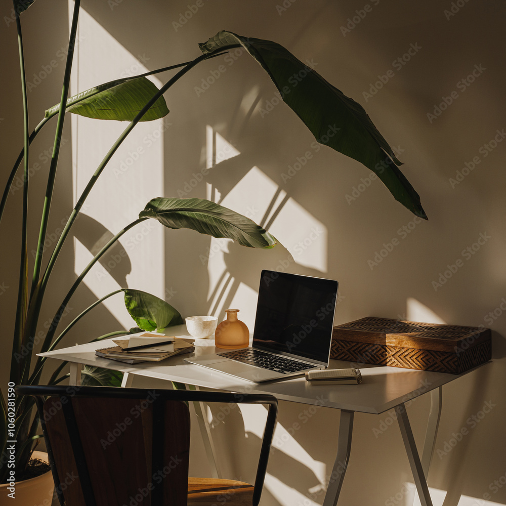 Naklejka premium Organized home workspace with stationery, laptop and tropical plant in a bright, airy room with aesthetic wam sunlight shadows