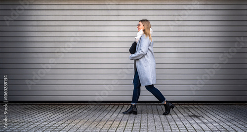 Middle-aged blonde woman in gray coat, scarf, jeans and sunglasses walking against background of gray modern wall in city on autumn day. Businesswoman. Side view. 