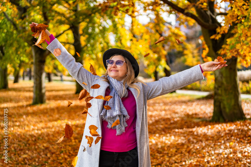 Portrait of happy middle-aged woman wearing gray coat and scarf, pink sweater, black brimmed hat throwing golden leaves against alley with autumn trees in background in city park on sunny day. Energy 