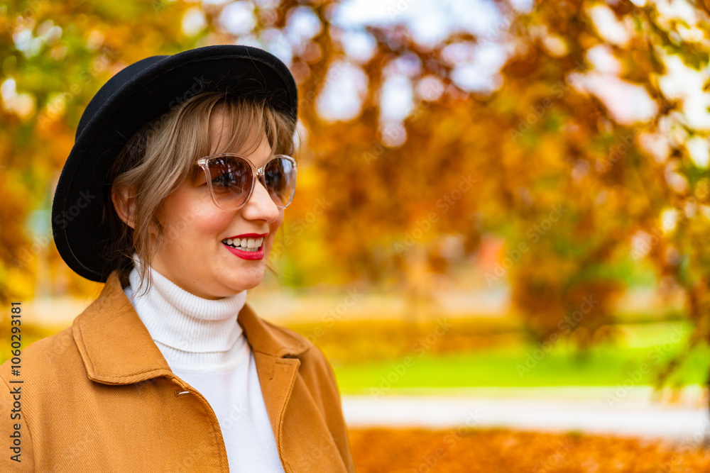 Portrait of beautiful young woman wearing beige coat, cream turtleneck, black brimmed hat and sunglasses standing in city park on autumn day.	