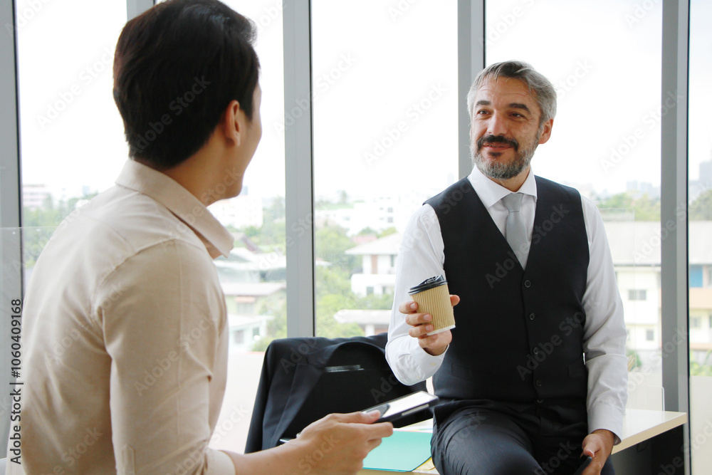 Smiling elderly senior Caucasian manager drinking coffee and talking with asian young businessman for brainstorming for project job together in office Smiling elderly senior Caucasian manager drinking coffee and talking with asian young businessman for brainstorming for project job together in office