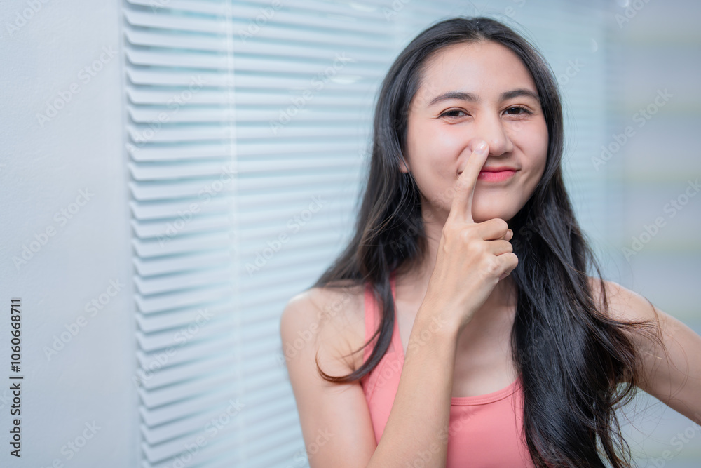 Young asian woman in pink top is touching her nose with her fingertip, expressing a playful and slightly mischievous attitude, against a background of venetian blinds Young asian woman in pink top is touching her nose with her fingertip, expressing a playful and slightly mischievous attitude, against a background of venetian blinds