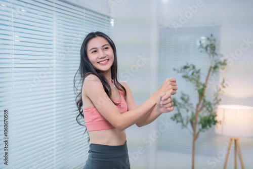Young asian woman wearing sportswear smiles while performing arm stretches in a bright room with window blinds and a small decorative tree Free Download Young asian woman wearing sportswear smiles while performing arm stretches in a bright room with window blinds and a small decorative tree - GFXLoad