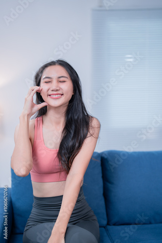 Happy asian fitness woman sitting on sofa and touching her face with closed eyes, enjoying the moment of relaxation after exercising at home