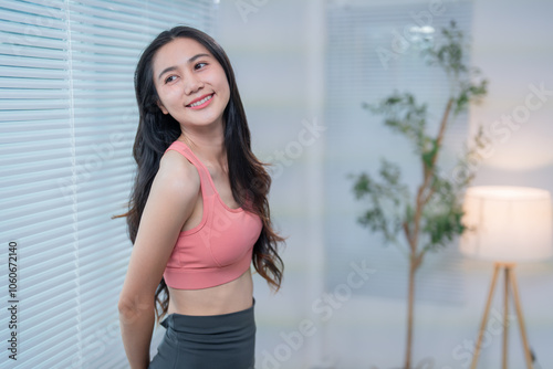 Fit asian woman wearing sportswear smiling and relaxing near window blinds at home after exercising, enjoying domestic leisure time