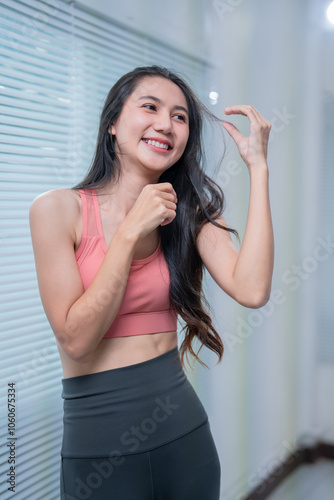 Happy sporty asian woman in sportswear touching her hair and smiling while standing near window blinds at home enjoying active healthy lifestyle