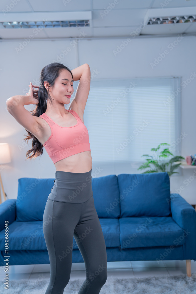 Fit asian woman in sportswear preparing for exercising at home, stretching her arms and tying her hair in a ponytail, with a blue sofa in the background Fit asian woman in sportswear preparing for exercising at home, stretching her arms and tying her hair in a ponytail, with a blue sofa in the background