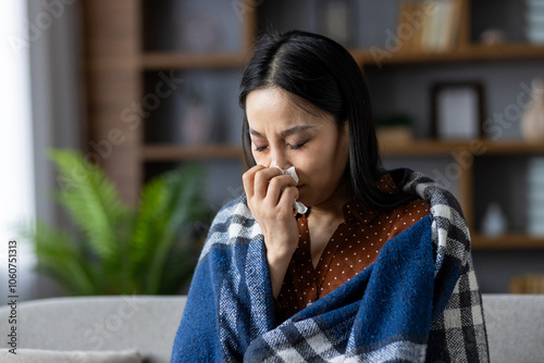 Asian woman sitting at home wrapped in warm blanket while holding tissue. She appears to be unwell, dealing with cold symptoms, seeking comfort and warmth indoors, feeling unwell.