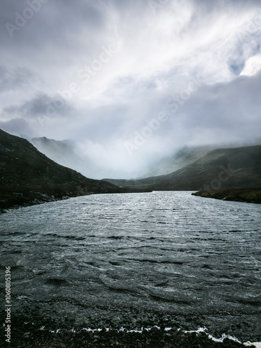 A calm river winds through a misty mountain valley, with fog rolling over the peaks. The water glimmers under a cloudy sky, creating a tranquil atmosphere. photo