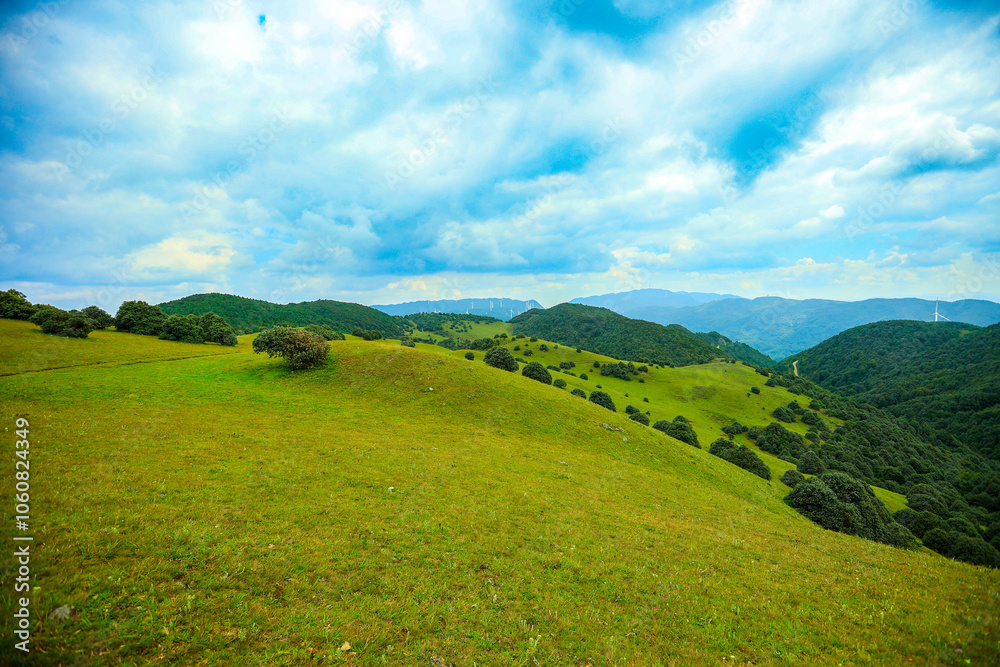 Fototapeta premium Serene Mountain Meadow with Distant Wind Turbines under Blue Sky 