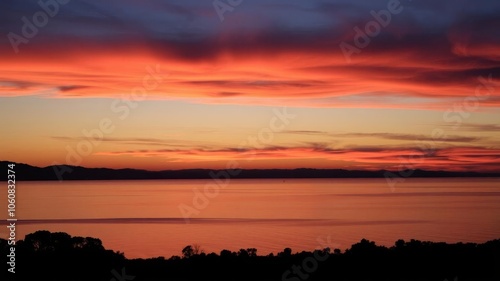 Vibrant orange and pink sunset reflecting on calm water with silhouettes of trees, sky, calm