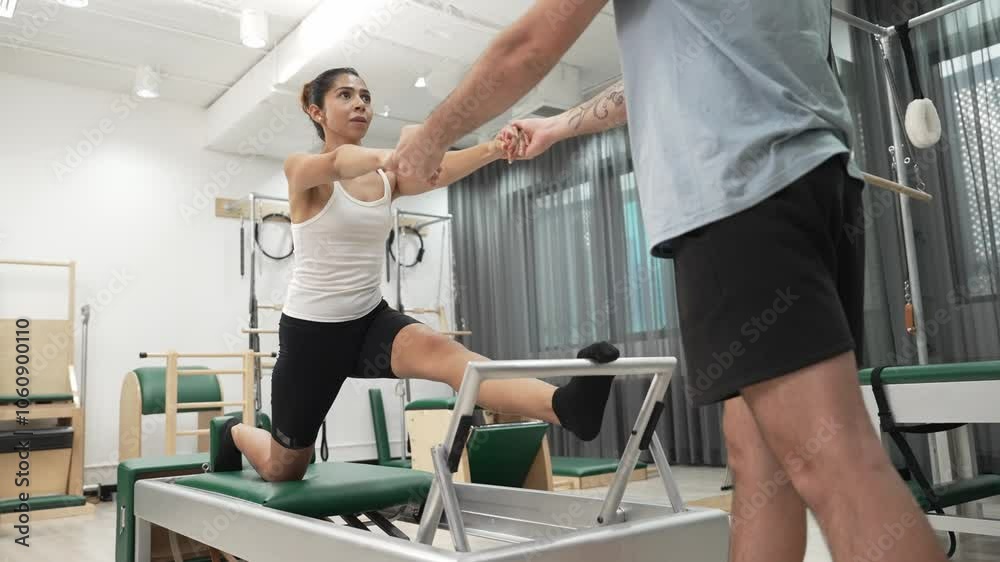 Woman receiving assistance in split stretch exercise during pilates class on trapeze table for flexibility and core training. Student doing split stretch on reformer for improving balance. Habituate. Woman receiving assistance in split stretch exercise during pilates class on trapeze table for flexibility and core training. Student doing split stretch on reformer for improving balance. Habituate.