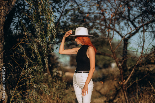 Red-haired lady wearing stylish white jeans and elegant black top, fashionable white hat posing gracefully next to tall tree, beautiful autumn scenery along picturesque riverside, colorful leaves scat photo