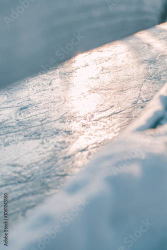 Close-up of smooth, sunlit ice surface on a skating rink with subtle grooves, creating a serene winter scene with soft light and frosty texture.
 photo