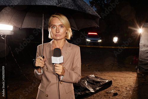 Waist up portrait of woman as news reporter standing at crime scene at night and looking at camera holding umbrella, copy space photo