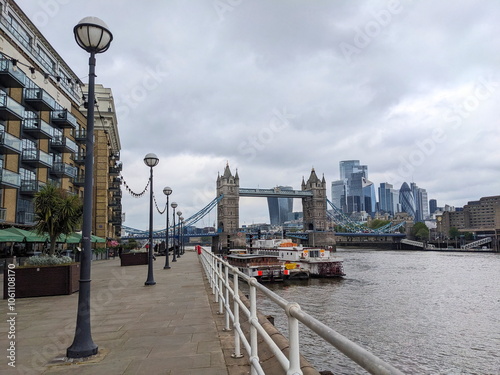 London, UK - 05.13.2024: A footpath on Butler's Wharf along outdoor seating of a restaurant and boats moored on the side of River Thames, with Tower Bridge and modern skyscrapers at the back