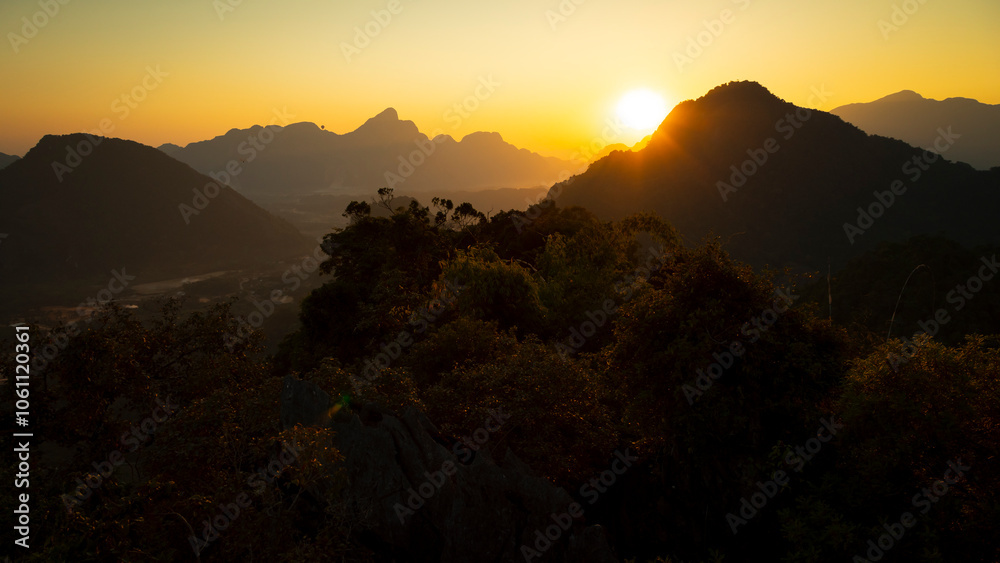 The last rays of sunshine pierce the warm air, turning the sky a fanciful orange and drawing the dark silhouettes of the mountains, Vang Vieng, Laos The last rays of sunshine pierce the warm air, turning the sky a fanciful orange and drawing the dark silhouettes of the mountains, Vang Vieng, Laos