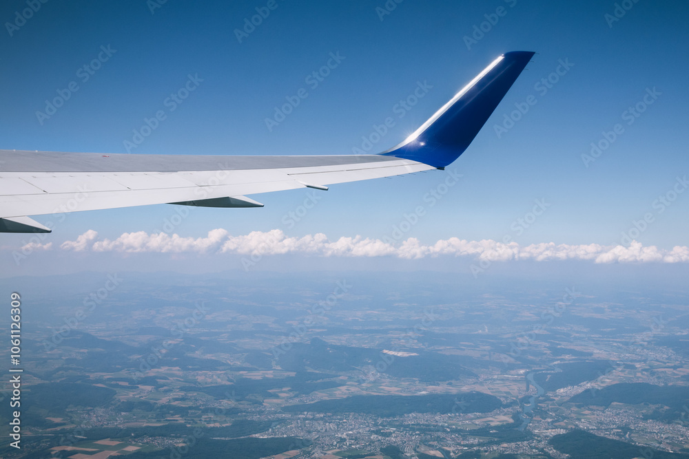 View from an airplane window of a wing against a clear blue sky, with clouds and a scenic landscape below. Captures the essence of air travel and aerial perspective. View from an airplane window of a wing against a clear blue sky, with clouds and a scenic landscape below. Captures the essence of air travel and aerial perspective.