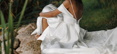 The afro bride sits gracefully on a rocky surface, her elegant white gown flowing around her as she embraces the calmness of a natural setting on her wedding day photo