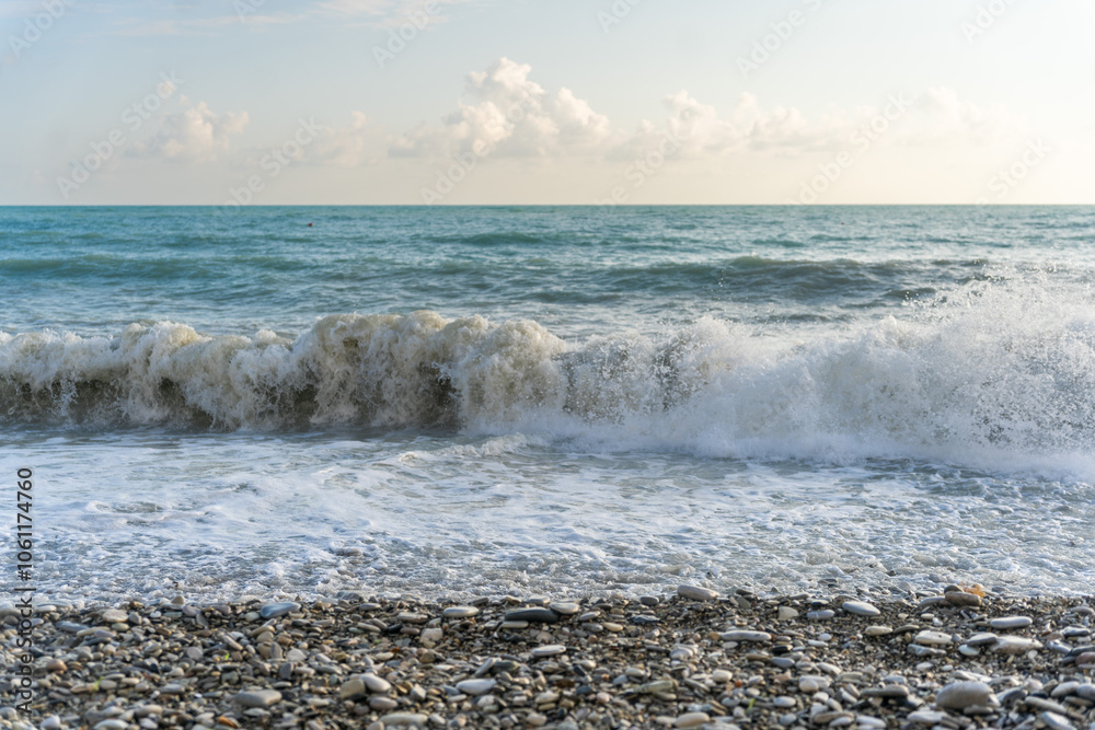 The sea waves roll onto a pebble sea shore against a blue sky with clouds. The sea waves roll onto a pebble sea shore against a blue sky with clouds.