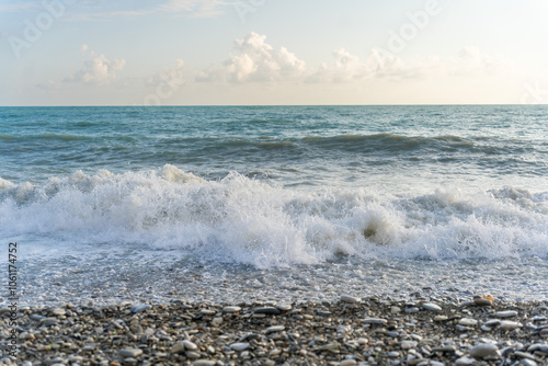 The sea waves roll onto a pebble sea shore against a blue sky with clouds.