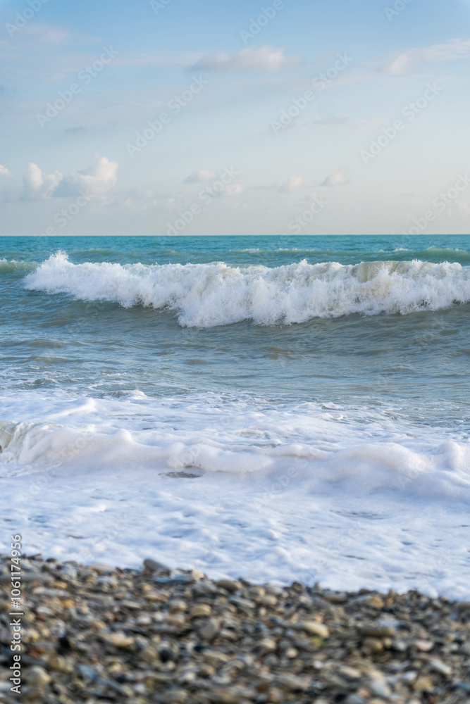The sea waves roll onto a pebble sea shore against a blue sky with clouds. The sea waves roll onto a pebble sea shore against a blue sky with clouds.