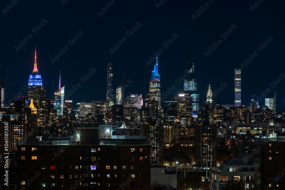 Fototapeta premium A stunning nighttime view of the New York City skyline with its iconic high-rise buildings illuminated in vibrant lights. The Empire State Building stands prominently with its colorful lights against 