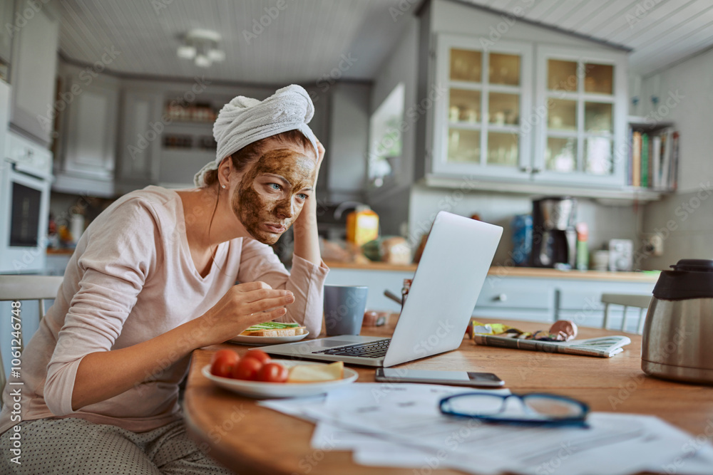 Woman with face mask working on laptop in kitchen during breakfast Woman with face mask working on laptop in kitchen during breakfast