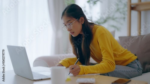 Young Asian female wearing glasses using laptop, working at home in living room, coffee mug on table. Cozy office workplace, remote work