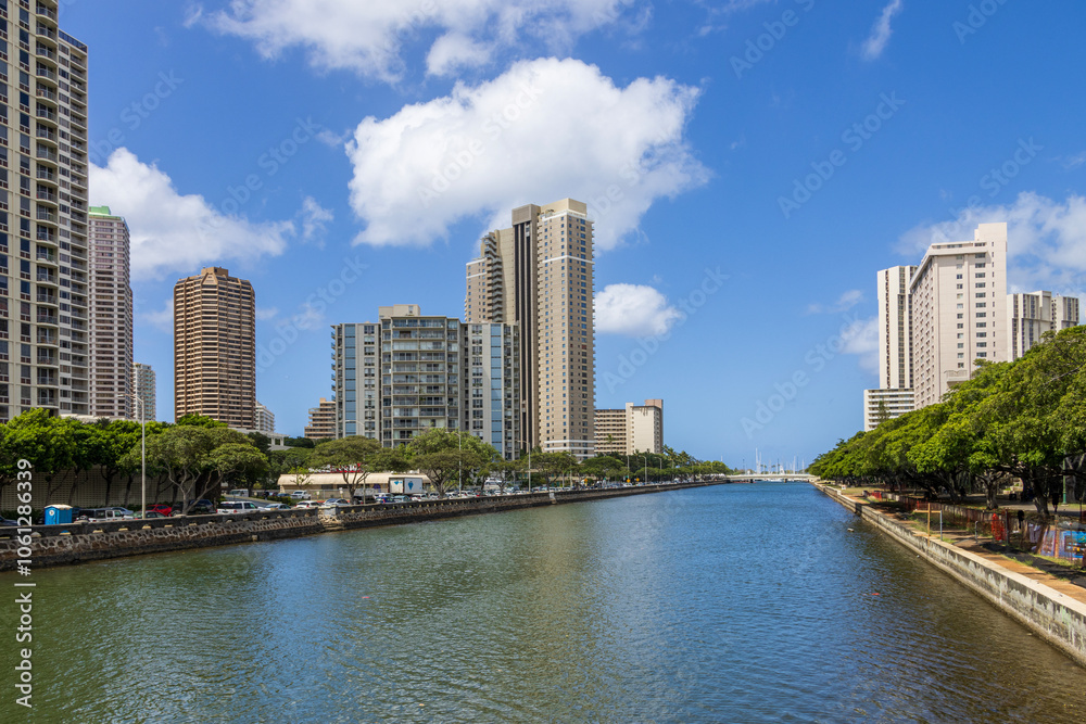 The rippling waters of the Ala Wai Canal with skyscrapers, hotels and condos, lush green trees, cars on the street, blue sky and clouds in downtown Honolulu Hawaii USA The rippling waters of the Ala Wai Canal with skyscrapers, hotels and condos, lush green trees, cars on the street, blue sky and clouds in downtown Honolulu Hawaii USA