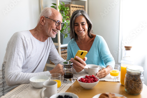 Happy mature caucasian married couple eating healthy breakfast at home looking at mobile phone.