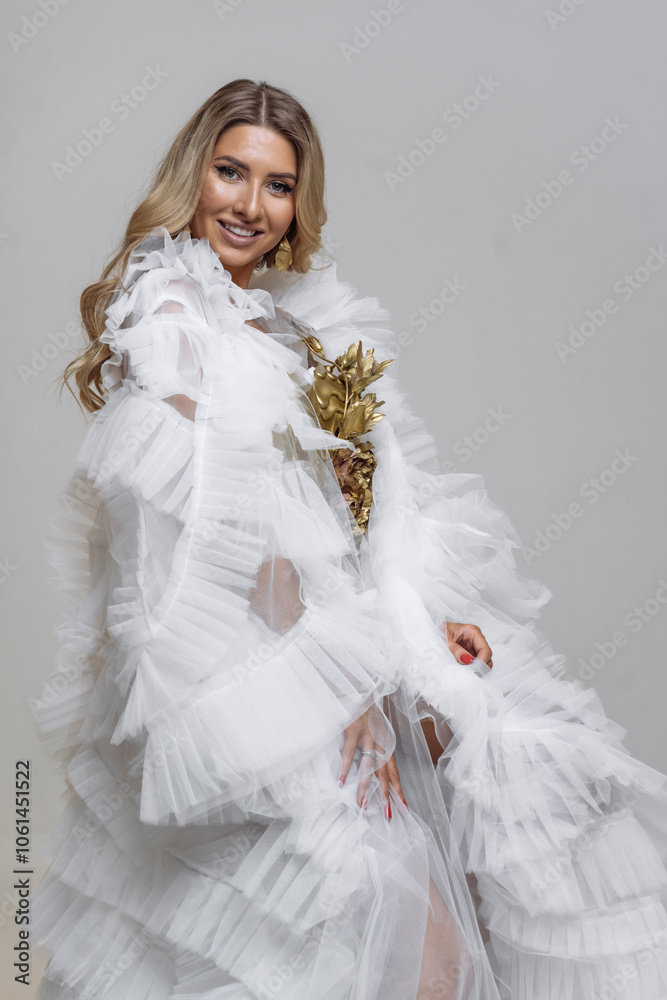 A smiling woman with long blonde hair poses in elegant outfit shewears a white ruffled dress with gold accents. A smiling woman with long blonde hair poses in elegant outfit shewears a white ruffled dress with gold accents.