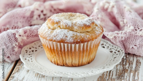 A freshly baked muffin dusted with powdered sugar sits elegantly on a delicate plate, surrounded by a soft, pink fabric.