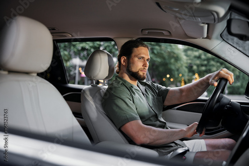 Man driving a car with a serious expression, focused on the road, interior view with blurred greenery in the background