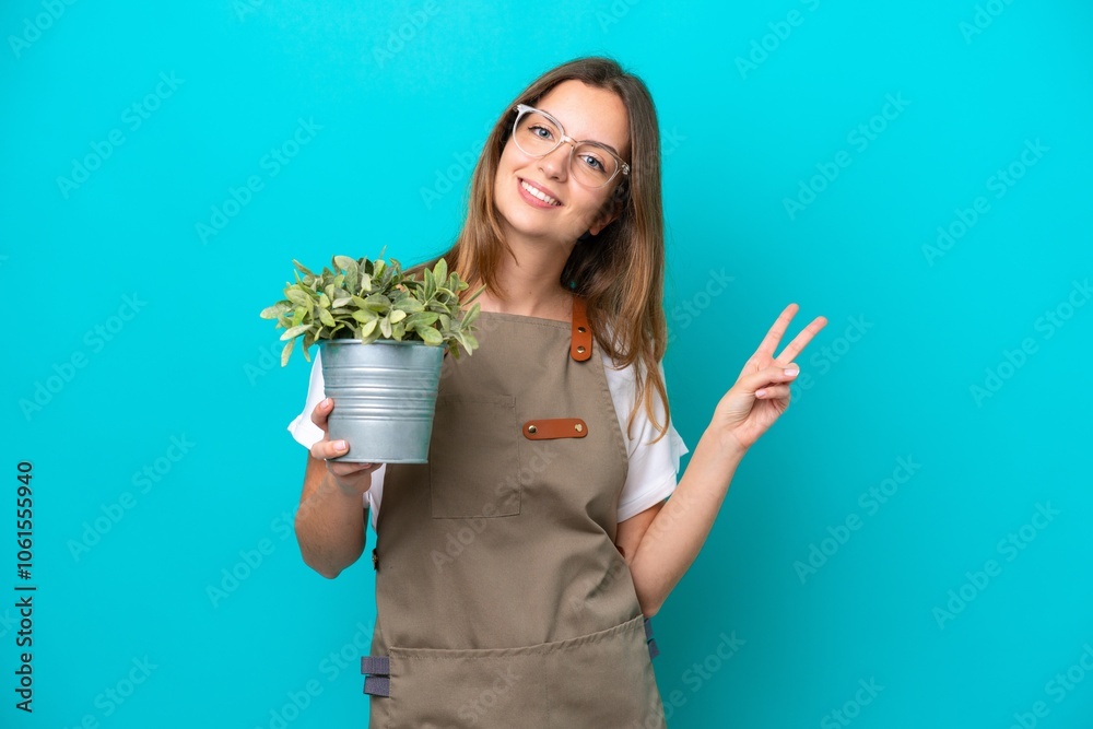 Young caucasian gardener woman holding a plant isolated on blue background smiling and showing victory sign Young caucasian gardener woman holding a plant isolated on blue background smiling and showing victory sign
