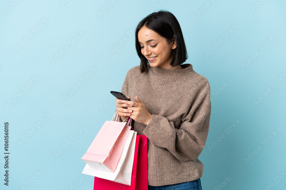 Young caucasian woman isolated on blue background holding shopping bags and writing a message with her cell phone to a friend Young caucasian woman isolated on blue background holding shopping bags and writing a message with her cell phone to a friend