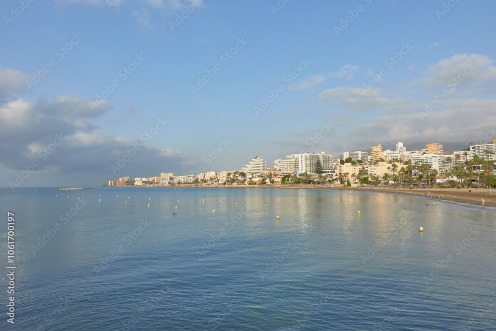 Naklejka premium Calm coastal cityscape of Benalmadena Costa, village of Malaga, Andalusia, Spain. Blue water and beachside buildings under a partly cloudy sky, showing a serene Mediterranean shoreline