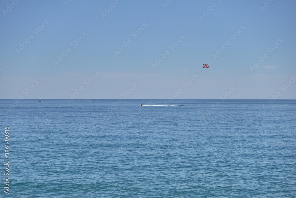 A serene seascape featuring calm blue waters under a clear sky, with a distant parasailing activity visible as a small red dot, creating a peaceful Mediterranean summer atmosphere A serene seascape featuring calm blue waters under a clear sky, with a distant parasailing activity visible as a small red dot, creating a peaceful Mediterranean summer atmosphere