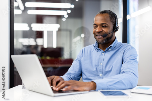 Mature african american businessman in modern office setting using laptop and headset. Displaying professionalism, communication skills, and confidence while typing and engaging with clients.