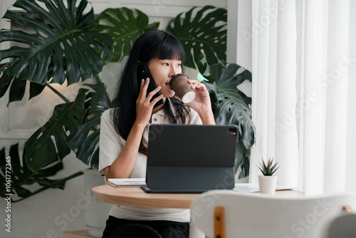 A young woman multitasks by talking on the phone and drinking coffee while working on her laptop in a modern home office with lush green plants. photo