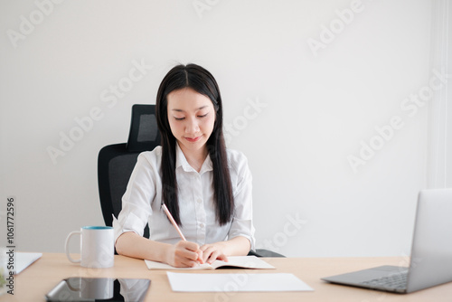 Young woman in a white shirt writing in a notebook at a desk with a laptop and coffee mug in a bright office.