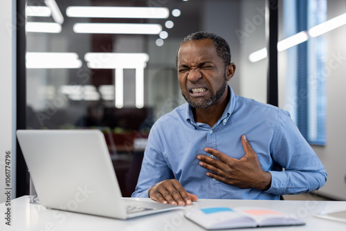 Mature businessman feeling chest pain at office desk, showing discomfort and stress. Conceptual image highlighting workplace health issues and emotional strain experienced by professionals.