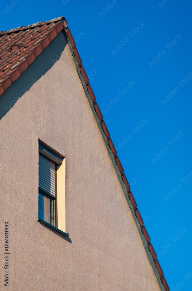 Obraz premium Gable end of house with clay roof tiles and window showing half-closed venetian blinds against blue sky