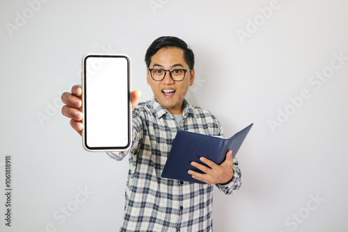 Asian college student holding book and blank smartphone screen with happy and excited expression. Young Asian student wearing blue shirt smiling and showing white mobile phone screen. photo