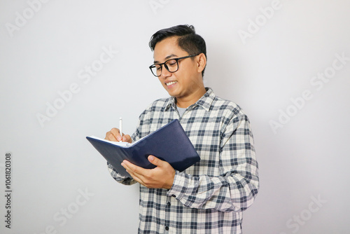 Professional and focused millennial Asian businessman in a formal business suit writing something on his personal planner book or schedule book stands on an isolated white background