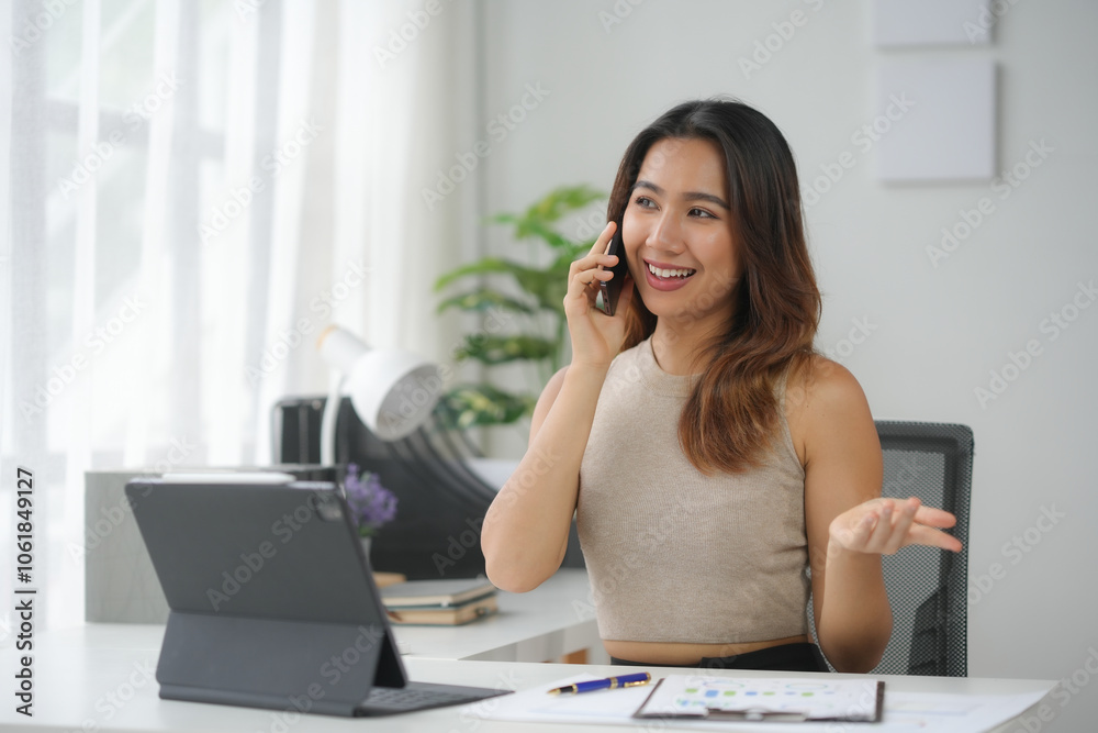 A smiling woman talking on the phone while working at her desk with a tablet and paperwork in a bright home office environment. A smiling woman talking on the phone while working at her desk with a tablet and paperwork in a bright home office environment.