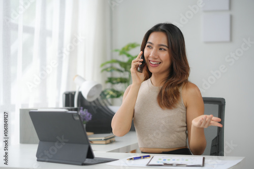 A smiling woman talking on the phone while working at her desk with a tablet and paperwork in a bright home office environment.