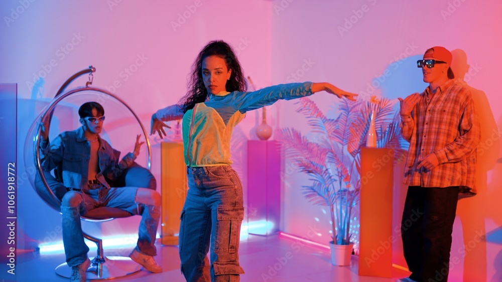 Fototapeta premium Attractive happy dancer looking at camera while smart woman moving at rhyme with neon light. Professional hispanic performer break dancing while wearing colorful cloth with diverse friend. Regalement.