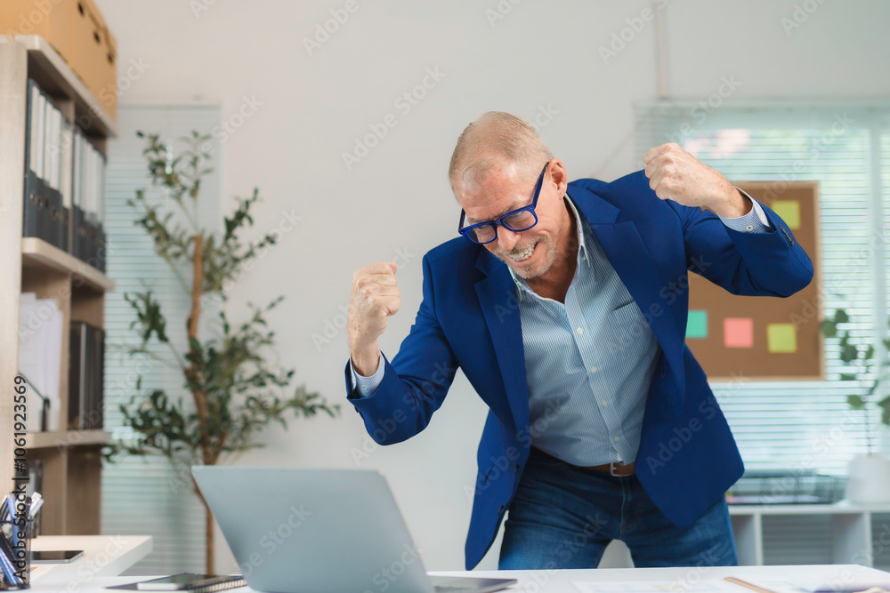 Fototapeta premium Senior businessman expressing excitement and joy while looking at his laptop in the office, celebrating a successful outcome