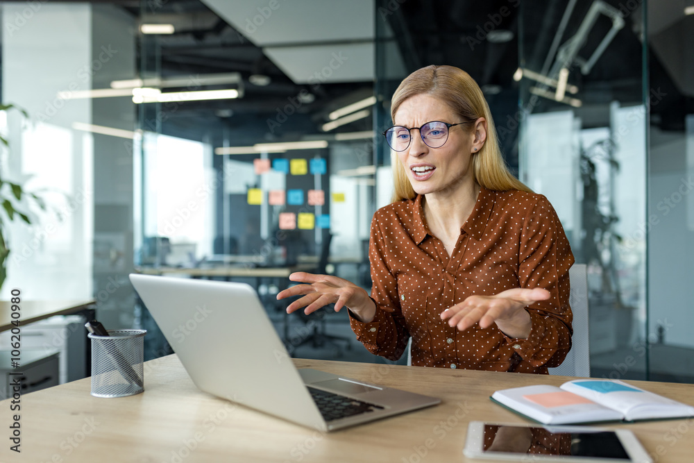© Liubomir - Mature business woman displays frustration during online conversation in office setting. Laptop and open notebook accentuate professional vibe, emphasizing challenges of remote communication.